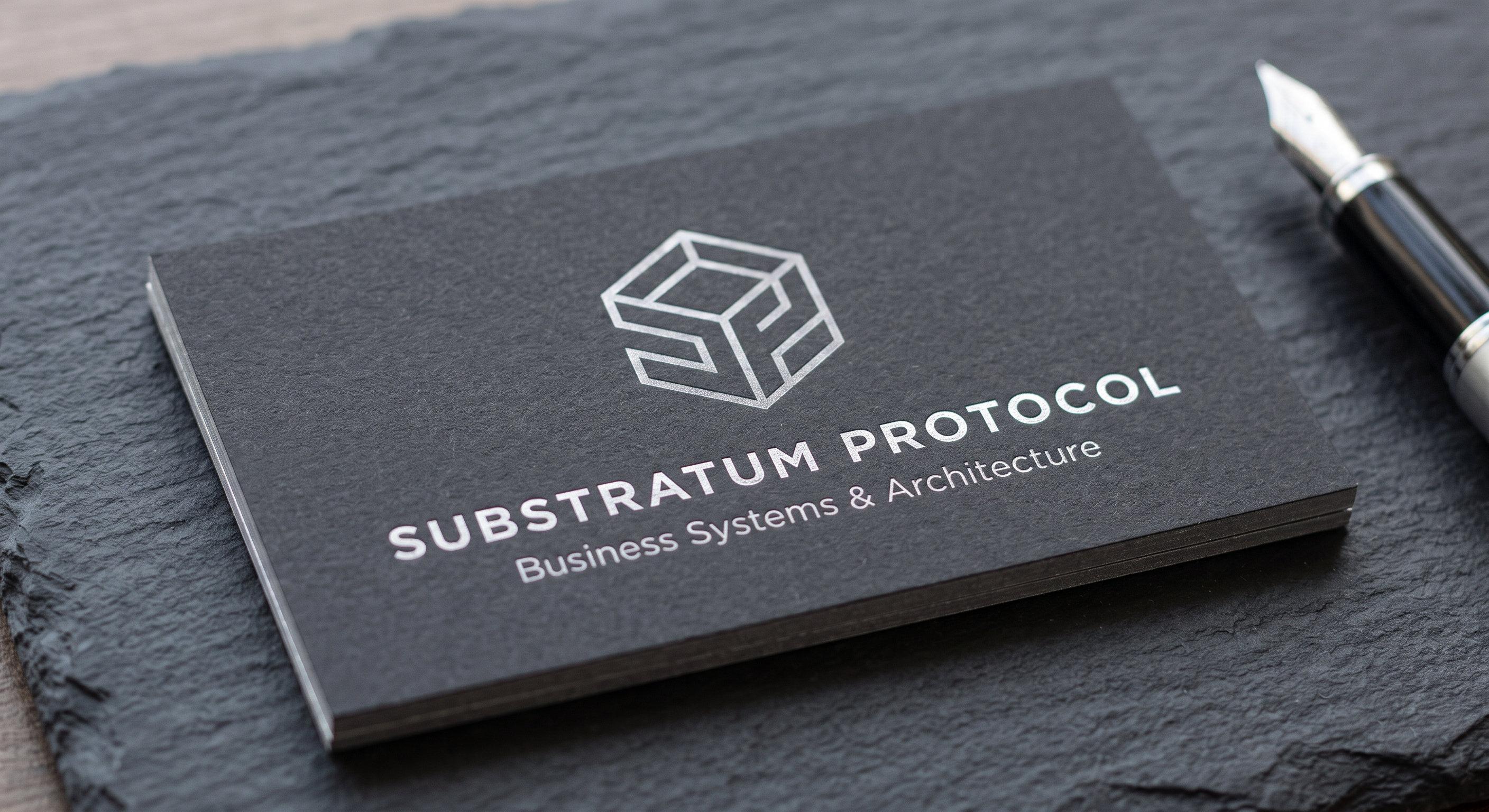 Stack of charcoal business cards with a silver-foil cube logo reading “Substratum Protocol — Business Systems & Architecture” beside a fountain pen